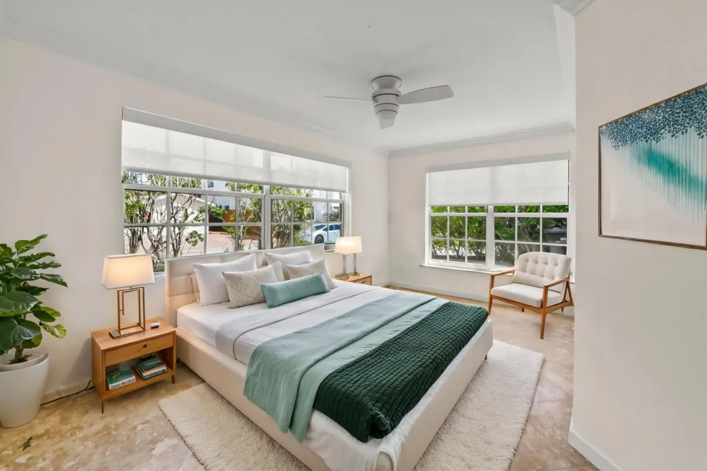Bright white bedroom with a low-profile bed, teal throw, two wooden nightstands, and large windows letting in natural light.