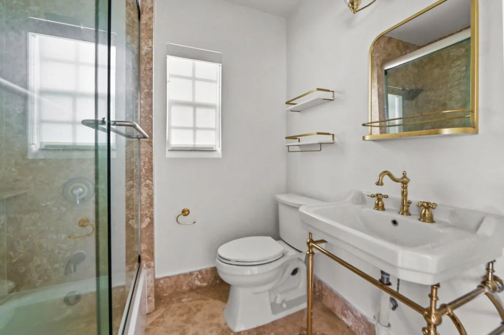 Modern bathroom with a glass shower enclosure, white toilet, marble floor, and gold fixtures under bright light.