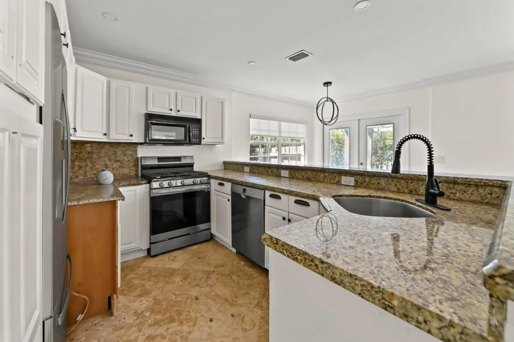Bright white kitchen with granite countertops, stainless steel appliances, and a central island sink with a black faucet.