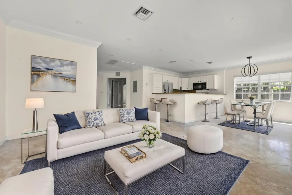 Open-concept living room with a white sofa, blue pillows, glass side table, and a white ottoman on a navy rug.