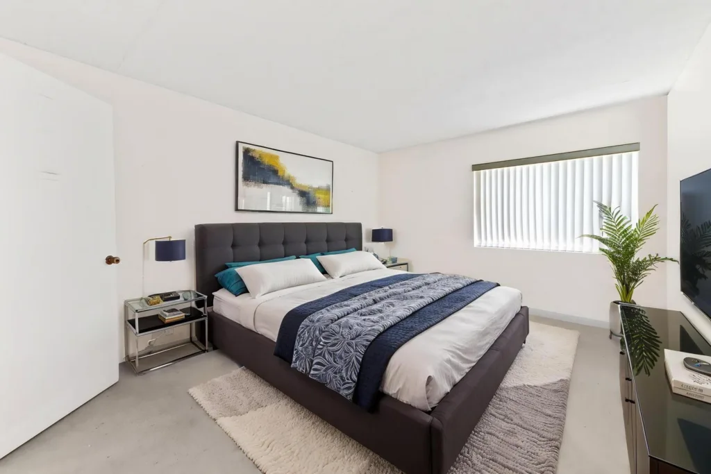 Modern bedroom with a large gray upholstered bed, white linens, and blue accents; abstract wall art above the headboard.