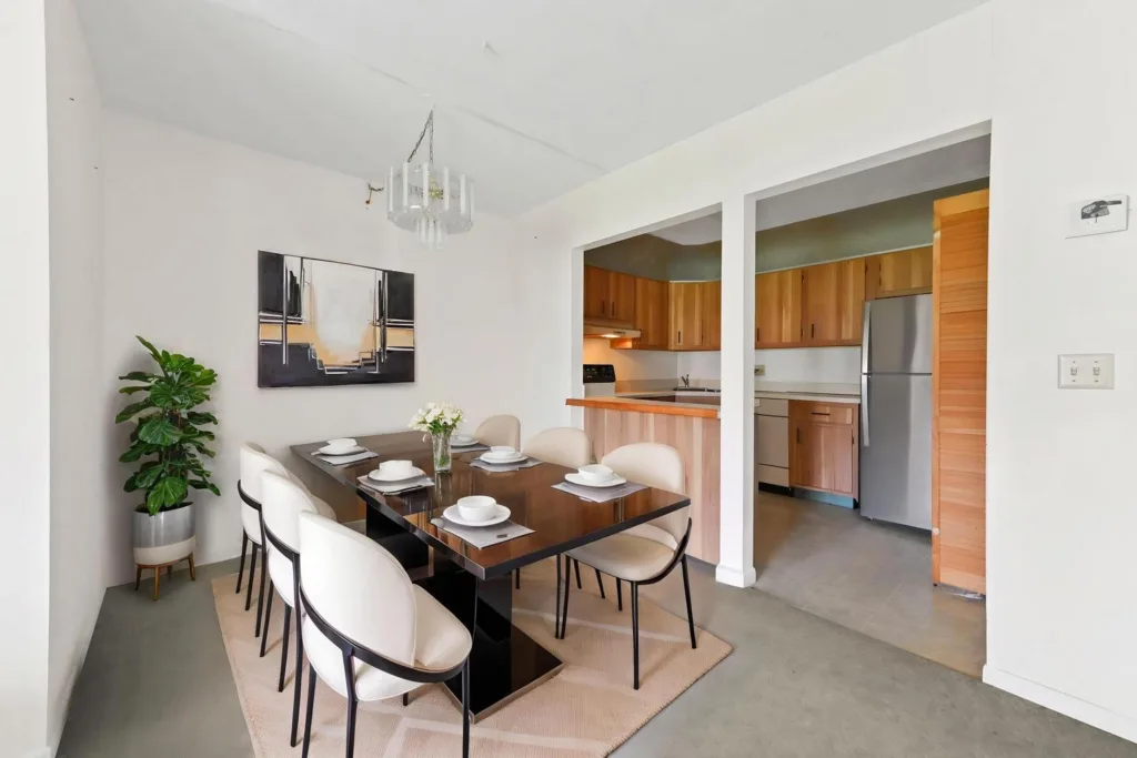 Modern dining room with a glass-top table set for six, cream chairs, and a chandelier, open to a wooden-cabinet kitchen.