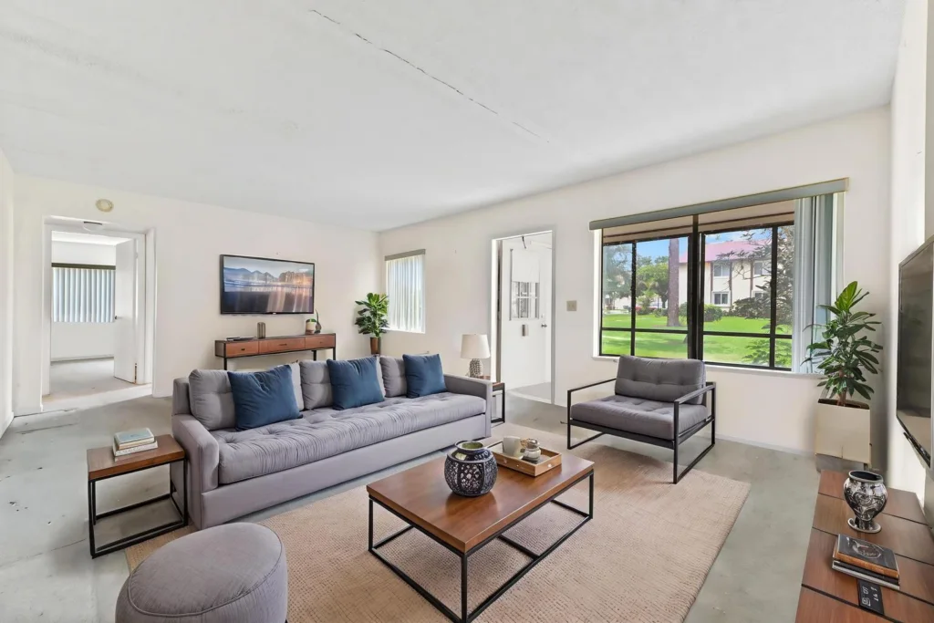 Bright living room with gray sectional sofa, coordinating chair, coffee table, TV, and large window view of greenery outside the room steadiness light.