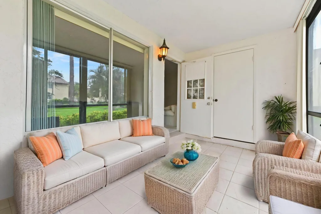 Cozy wicker seating area on a screened patio with beige cushions, orange and blue pillows, and a woven coffee table with a teal vase and pastries.