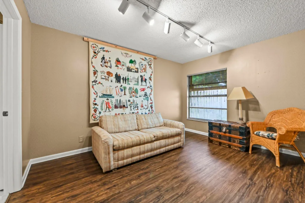 20261002200128980108000000-o - Lake Worth Homes - Lake Worth Homes Living room with beige sofa, wicker chair, and a large decorative wall hanging; wood-like flooring and track lighting above.