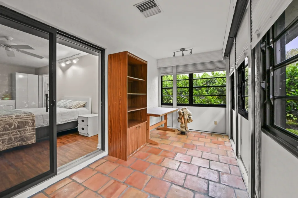 20261002200116548496000000-o - Lake Worth Homes - Lake Worth Homes Sunlit enclosed sunroom with terracotta tile, a tall wooden cabinet, a small desk, and a decorative dog statue, overlooking green shrubbery outside the windows.
