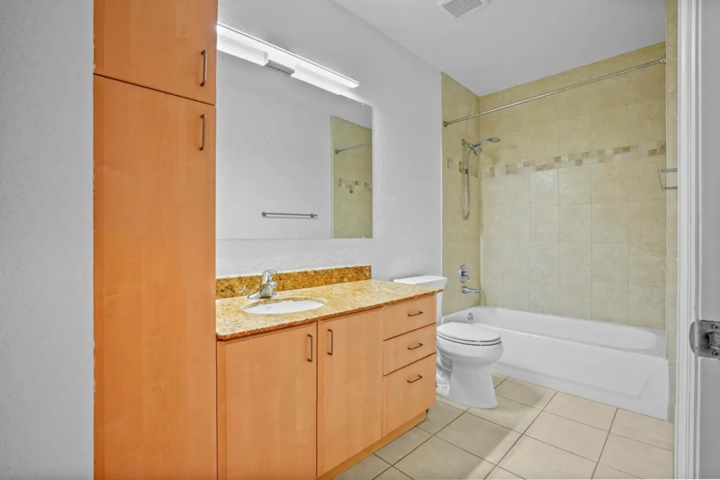 20261002144432927470000000-o - Lake Worth Homes - Lake Worth Homes Wood vanity with granite countertop and oval sink in a bright bathroom; mirror above and cabinet storage to the side; separated shower/tub with beige tiles.