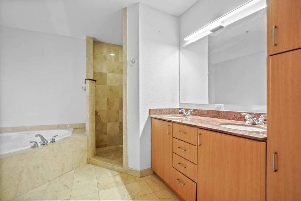 20261002144359713038000000-o - Lake Worth Homes - Lake Worth Homes Bathroom with a double-sink vanity, marble countertop, and wooden cabinets; beige-tiled tub and separate shower area to the left.