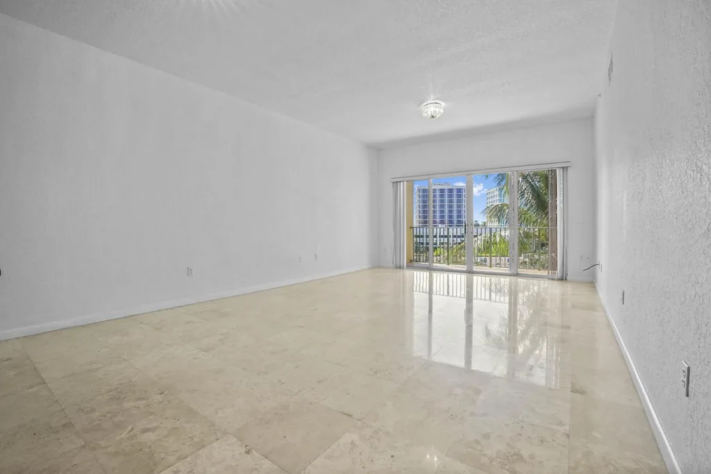 20261002144332832237000000-o 1 - Lake Worth Homes - Lake Worth Homes Empty white living room with glossy tile floor and sliding glass doors to a balcony overlooking palm trees and a cityscape outside.