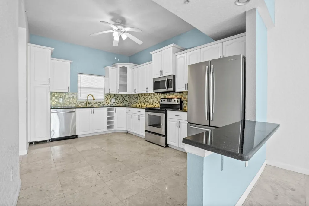 20261002144320100188000000-o 1 - Lake Worth Homes - Lake Worth Homes Bright L-shaped kitchen with white cabinets, stainless steel refrigerator, stove, and dishwasher, blue walls, mosaic tile backsplash, and beige tiled floor.
