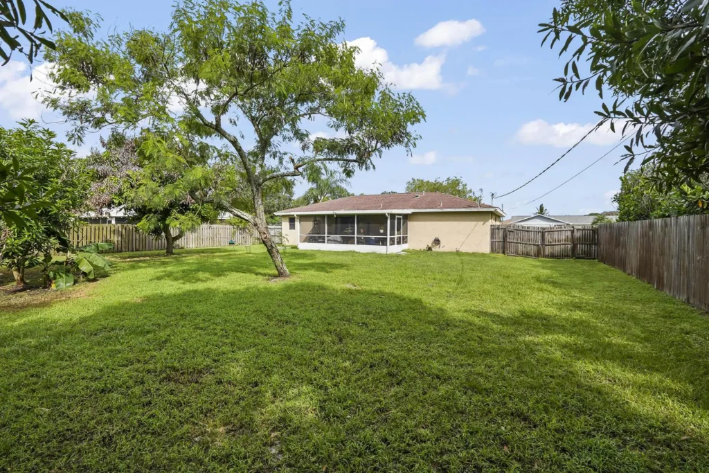 20261008152638316396000000-o - Lake Worth Homes - Lake Worth Homes Backyard of a single-story beige house with a screened porch, green lawn, and trees, fenced on all sides.