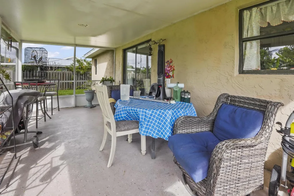 20261008152606279758000000-o - Lake Worth Homes - Lake Worth Homes Covered patio with a blue checkered tablecloth table and wicker chairs, overlooking a fenced backyard garden.