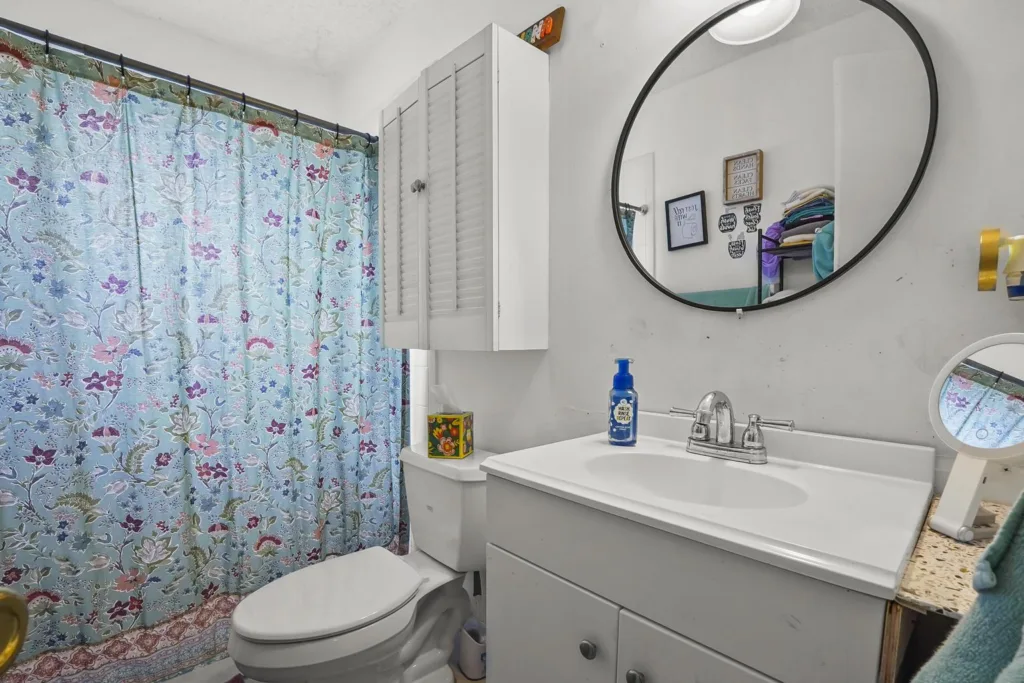 20261008152554060240000000-o - Lake Worth Homes - Lake Worth Homes Bathroom with a blue floral shower curtain, white toilet, and white vanity with round mirror above the sink.
