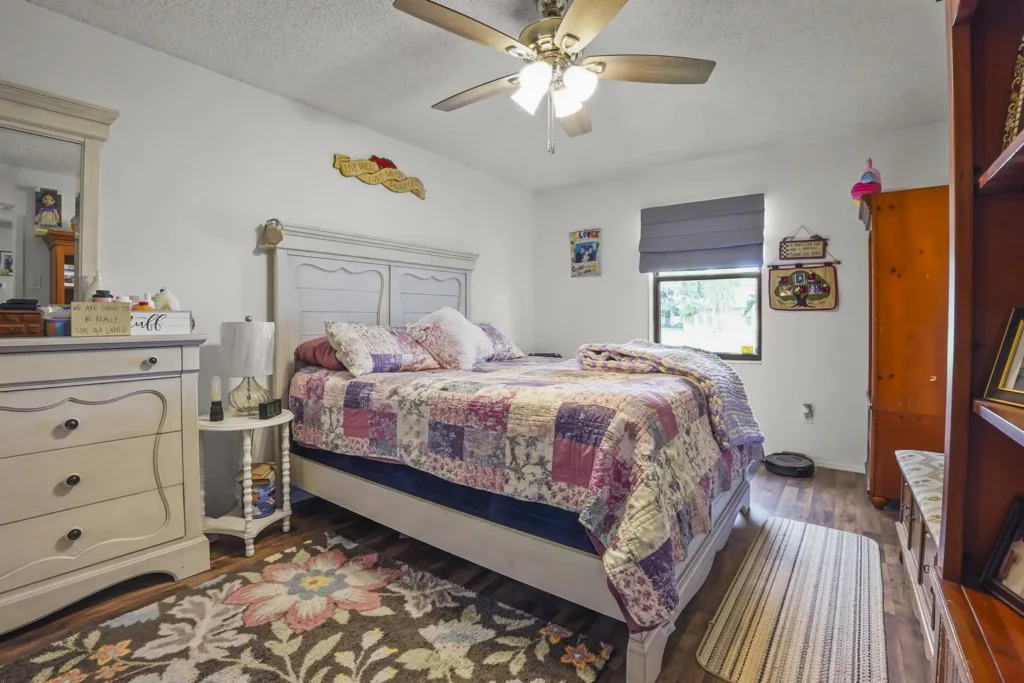 20261008152520951282000000-o - Lake Worth Homes - Lake Worth Homes Cozy bedroom with a white bed frame and a patchwork quilt, flanked by a dresser and nightstand under a ceiling fan.