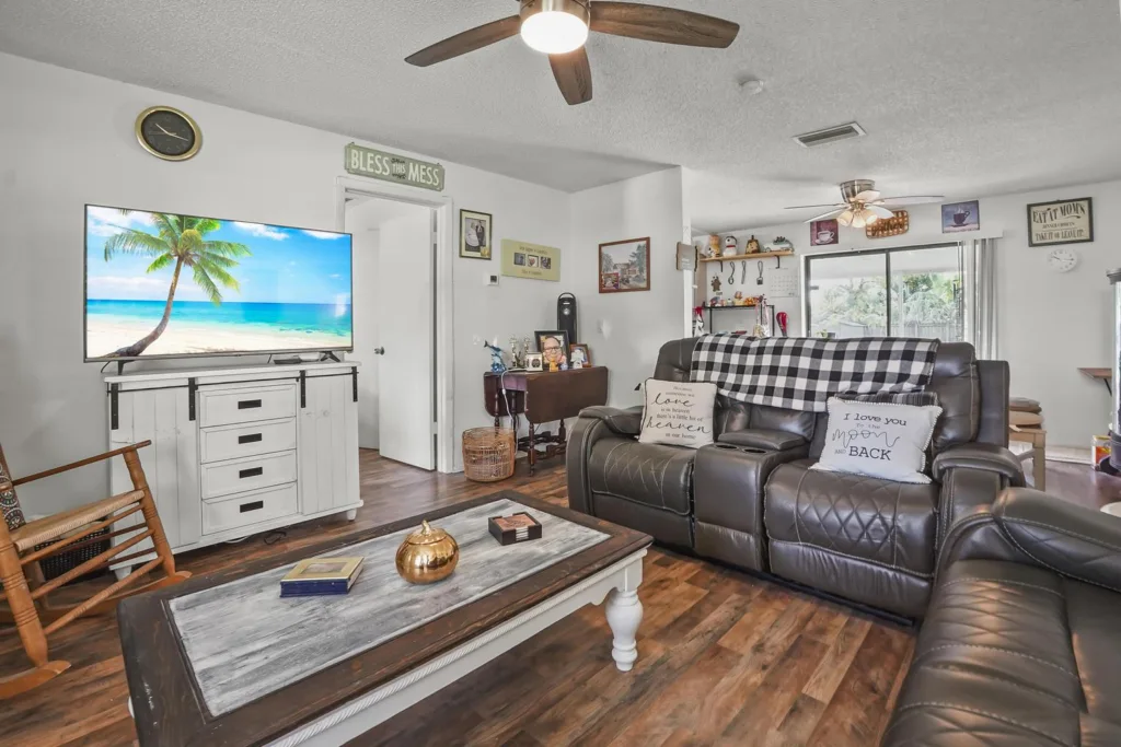 20261008152240876731000000-o - Lake Worth Homes - Lake Worth Homes Living room with a large flat-screen TV on a white cabinet, showing a tropical beach scene, and a dark leather sectional sofa nearby.