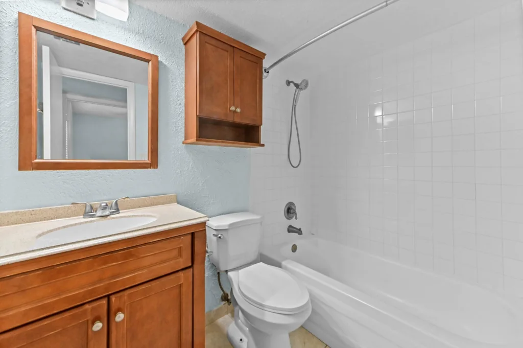 20261007172217378378000000-o - Lake Worth Homes - Lake Worth Homes Bathroom with a wooden vanity, wall mirror, and wall cabinet, next to a toilet and a white tiled bathtub/shower area