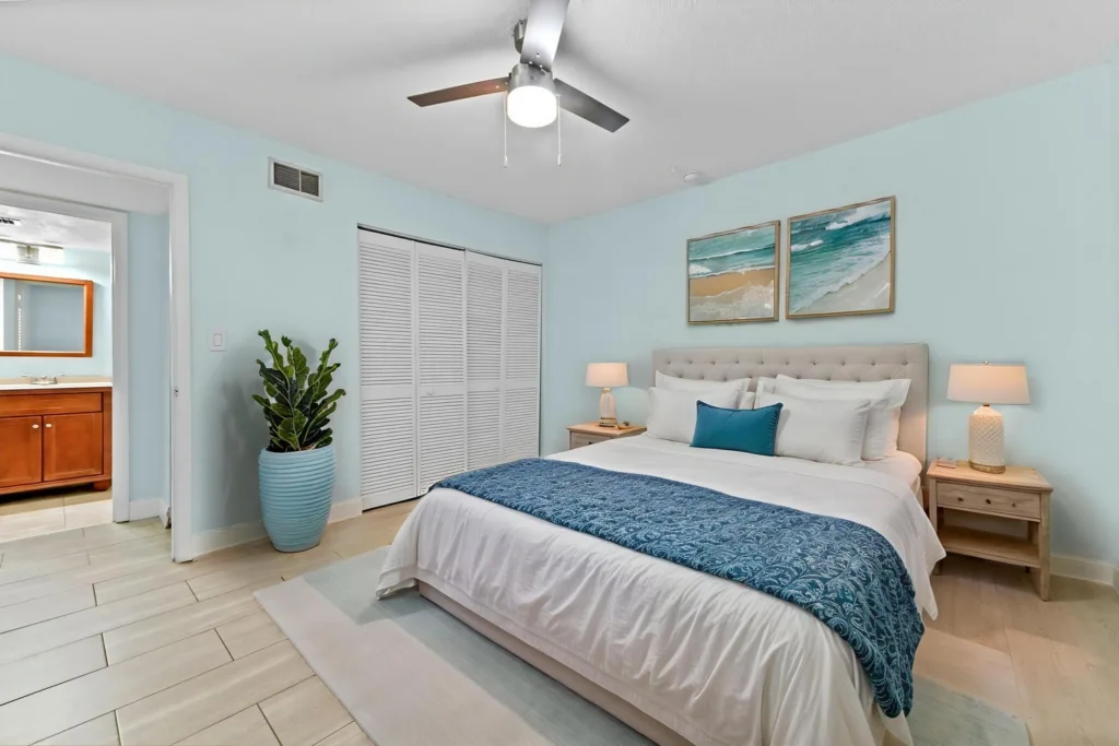 20261007173427470078000000-o - Lake Worth Homes - Lake Worth Homes Coastal bedroom with a queen bed, tufted headboard, blue throw, and two nightstands with lamps; beach artwork above the bed and a large potted plant nearby.