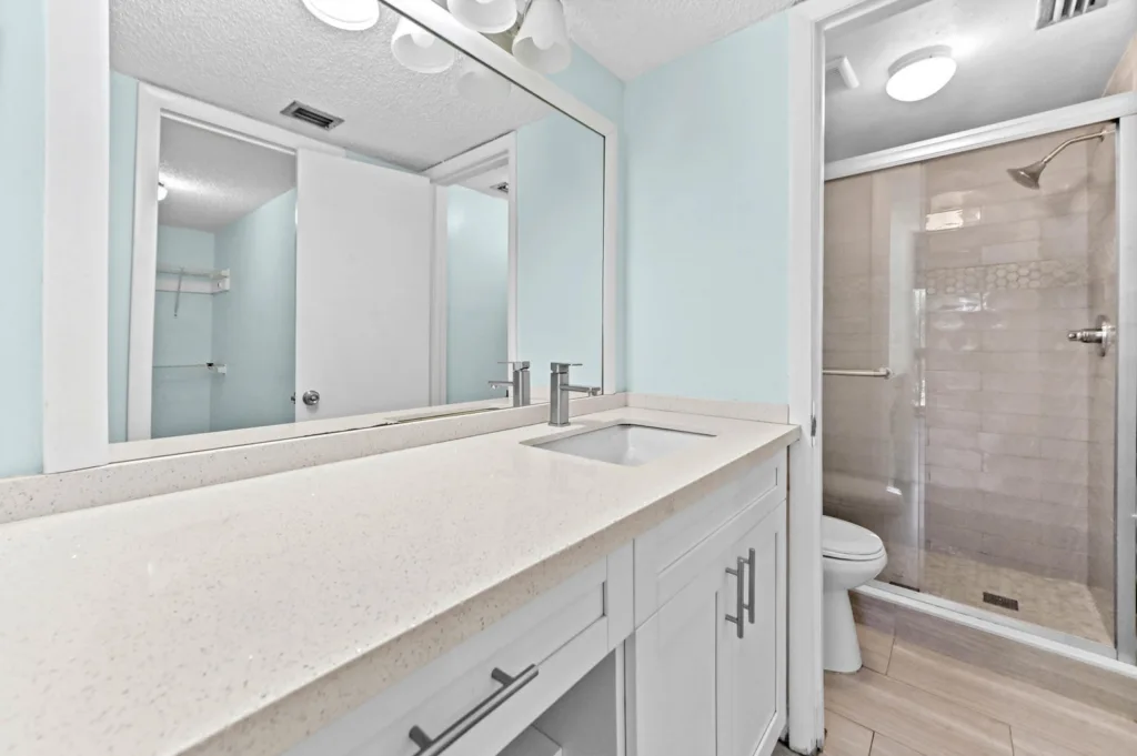 20261007172250198397000000-o - Lake Worth Homes - Lake Worth Homes White bathroom vanity with double sinks, a large mirror, light blue walls, and a glass-enclosed shower.