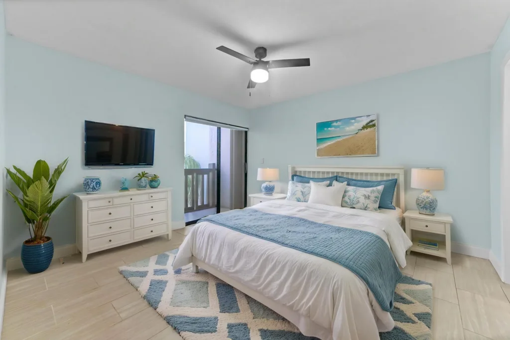 20261007173432023284000000-o - Lake Worth Homes - Lake Worth Homes Coastal-style bedroom with a white bed, blue quilt and pillows, white nightstands, lamps, and beach artwork above the headboard.
