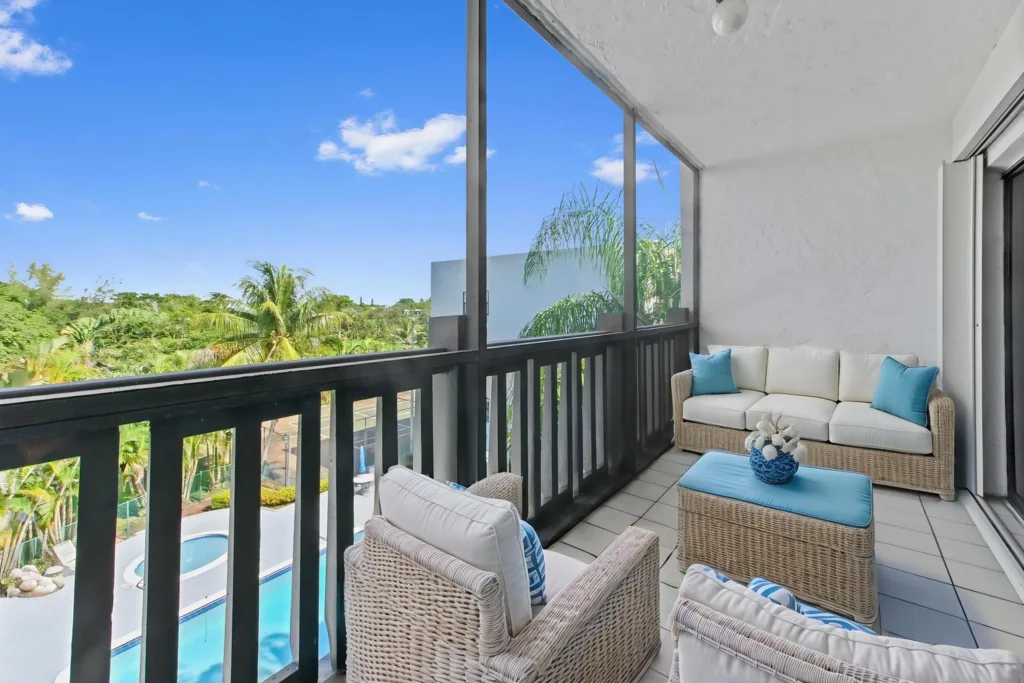 20261007173437590731000000-o - Lake Worth Homes - Lake Worth Homes Screened balcony with wicker sofa and chairs, light cushions, and turquoise pillows overlooking a palm-lined outdoor area and pool.