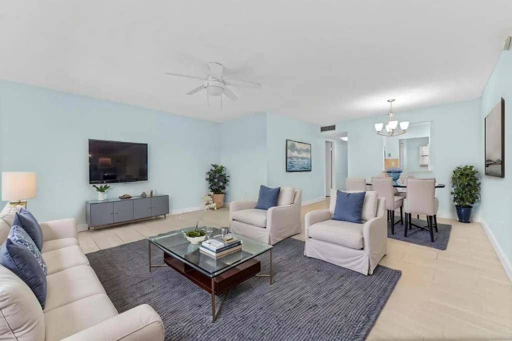 20261007173423167159000000-o - Lake Worth Homes - Lake Worth Homes Living room with light blue walls, beige sofas, and a glass coffee table on a dark rug; wall TV and modern console nearby.