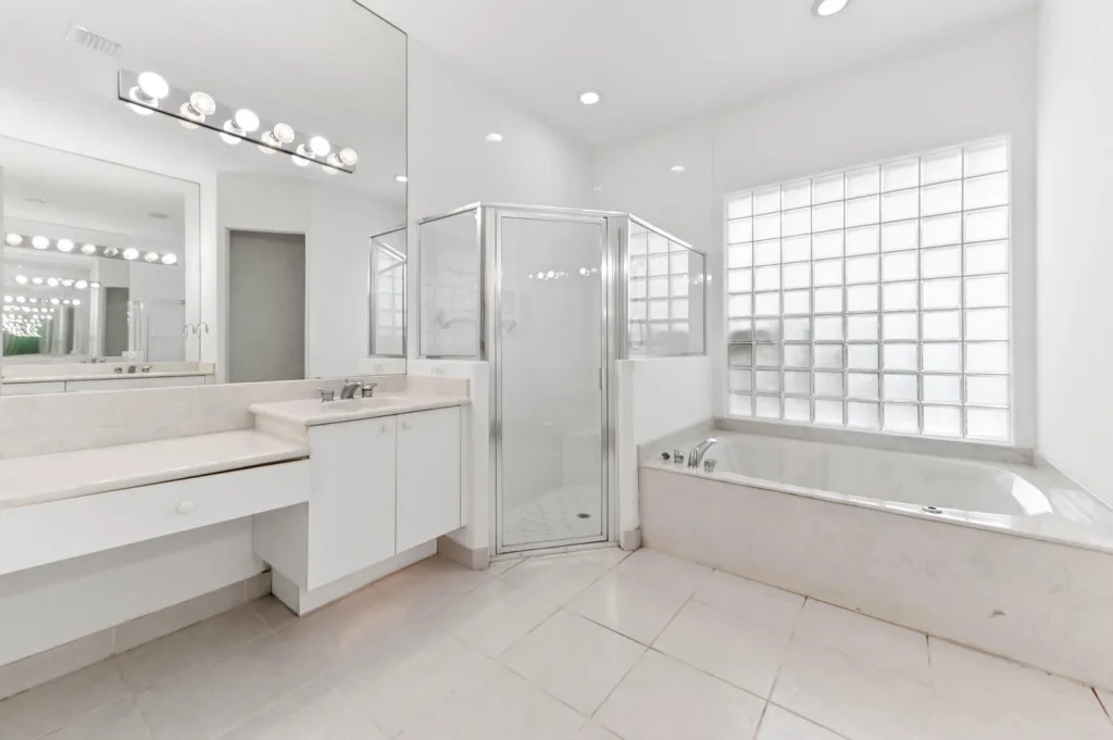 20261007212610718562000000-o - Lake Worth Homes - Lake Worth Homes Bright white bathroom with a double-sink vanity, glass-enclosed shower, and corner soaking tub near a glass-block window.