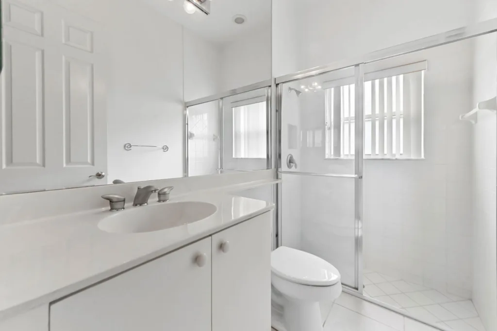 20261007212518703322000000-o - Lake Worth Homes - Lake Worth Homes Bright white bathroom featuring a single-sink vanity, large mirror, toilet, and glass sliding-shower enclosure with chrome hardware