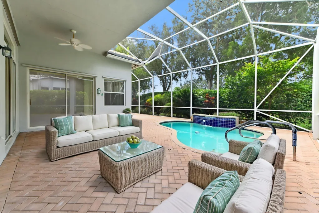 20261007221739334422000000-o - Lake Worth Homes - Lake Worth Homes Wicker sectional seating with cream cushions and teal pillows on a brick patio, facing a curvy turquoise pool inside a glass-enclosed patio.