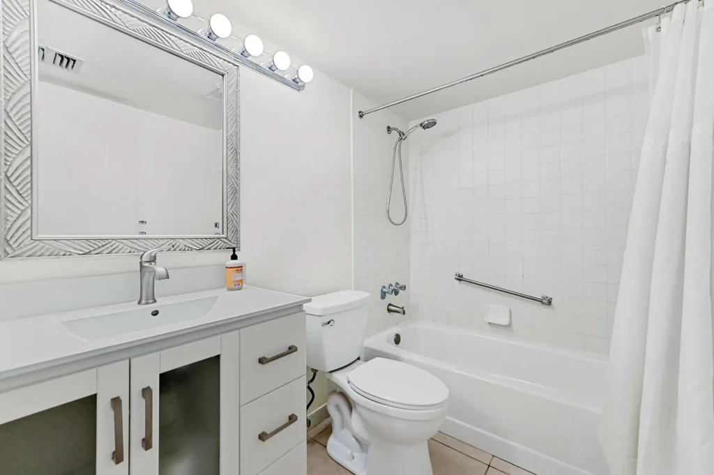 20261007235033201509000000-o - Lake Worth Homes - Lake Worth Homes White bathroom featuring a vanity with sink, decorative framed mirror, row of globe vanity lights, toilet, and a tiled bathtub/shower with a showerhead, grab bar, and white curtain.