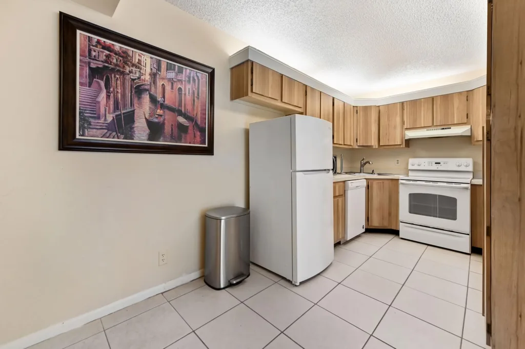 20261007234932741955000000-o - Lake Worth Homes - Lake Worth Homes Kitchen with white fridge, stove, dishwasher, and wooden cabinets; a framed canal scene hangs on the beige wall above a stainless trash can.