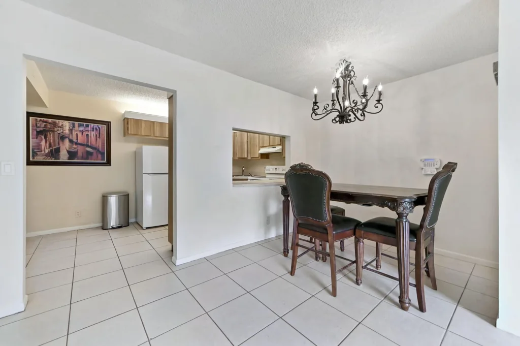 20261007234922994669000000-o - Lake Worth Homes - Lake Worth Homes Dining area with a dark wood table and four upholstered chairs under an ornate chandelier; kitchen pass-through visible in the background.