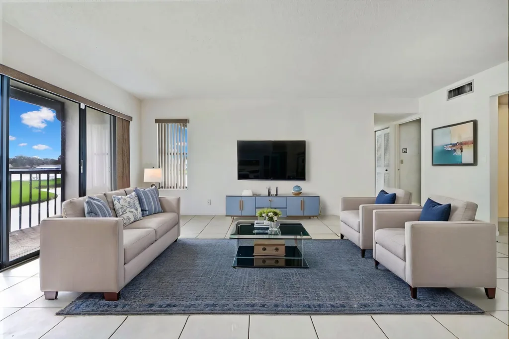 20261007234842393413000000-o 1 - Lake Worth Homes - Lake Worth Homes Bright living room with beige sofas, blue throw pillows, and a glass coffee table on a blue rug under a wall-mounted TV.
