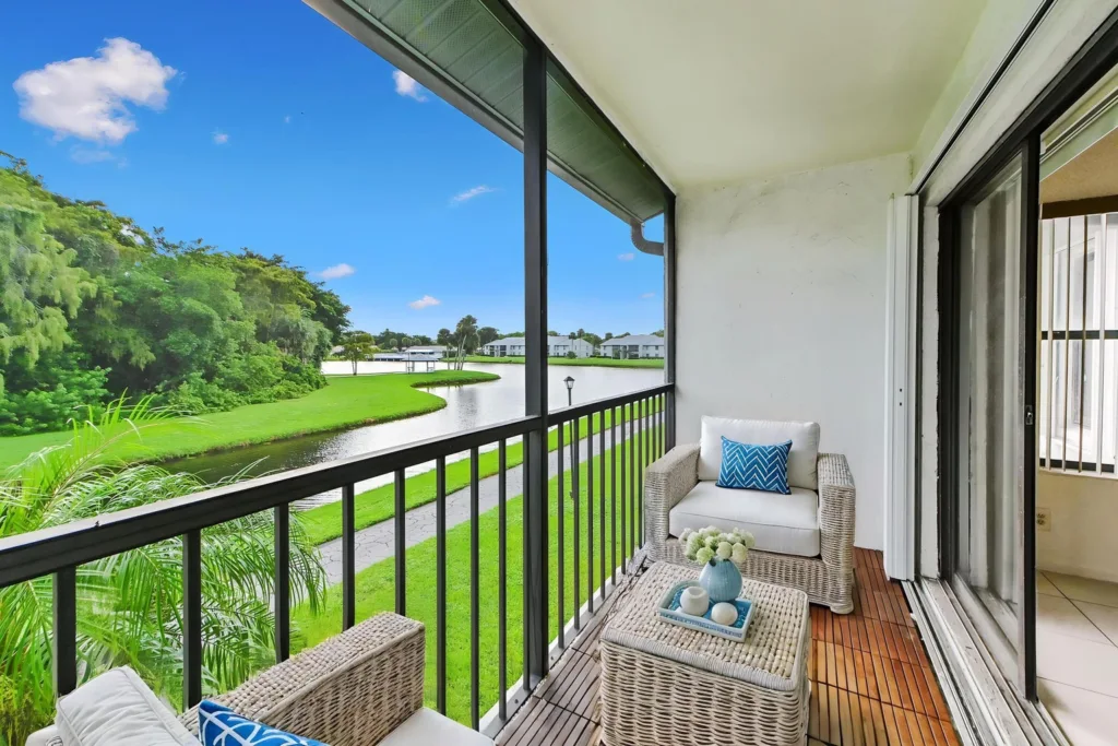 20261007235157402442000000-o 1 - Lake Worth Homes - Lake Worth Homes Balcony seating area with white wicker chairs and a wicker coffee table, overlooking a lake and green lawn on a sunny day.
