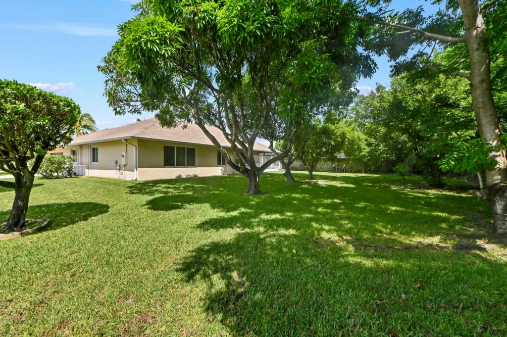20261006194042881130000000-o - Lake Worth Homes - Lake Worth Homes A sunny suburban backyard with several green trees and a beige single-story house in the background, casting shadows on a well-kept lawn.