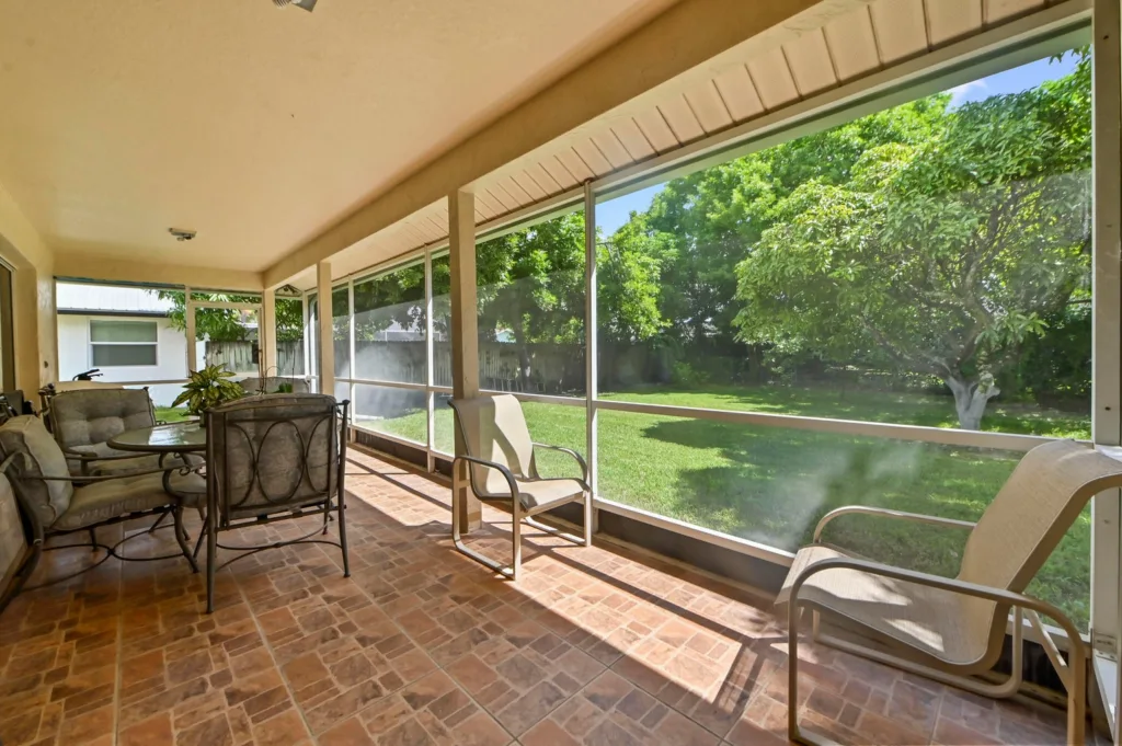 20261006193910116481000000-o - Lake Worth Homes - Lake Worth Homes Enclosed porch with brick-tiled floor, seating for six, and a view of a green backyard through screened walls that block the wind.