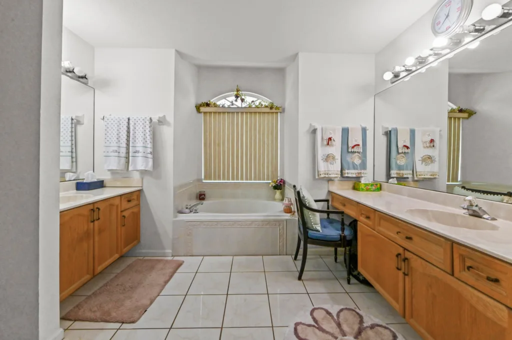20261006193845030921000000-o - Lake Worth Homes - Lake Worth Homes Bright bathroom with two wooden vanities, large mirror, and double sinks; towels hung on racks, bathtub under arched window with vertical blinds, and a blue chair nearby.