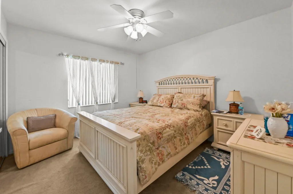 20261006193735376675000000-o - Lake Worth Homes - Lake Worth Homes Cream-colored bedroom with a floral quilted bed, lattice headboard, and matching nightstands on either side under a ceiling fan.