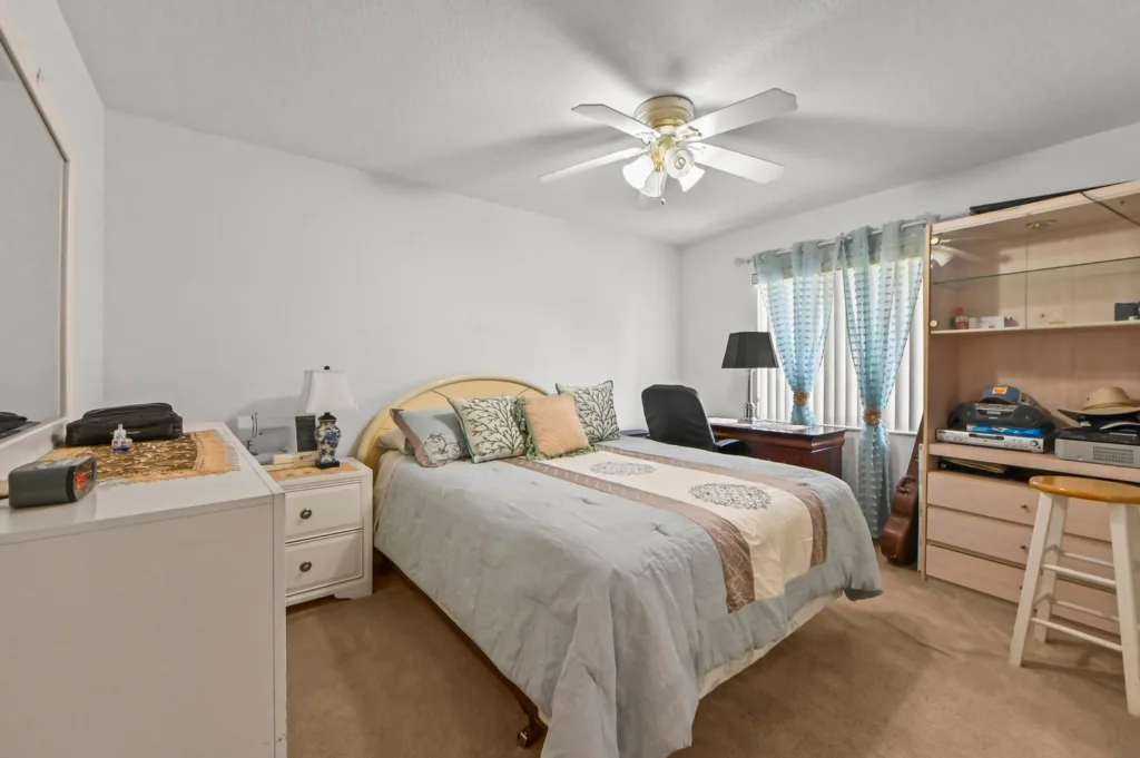 20261006193710587845000000-o - Lake Worth Homes - Lake Worth Homes A cozy bedroom with a queen bed, light blue comforter, and decorative pillows, a white dresser to the left, and a ceiling fan above.