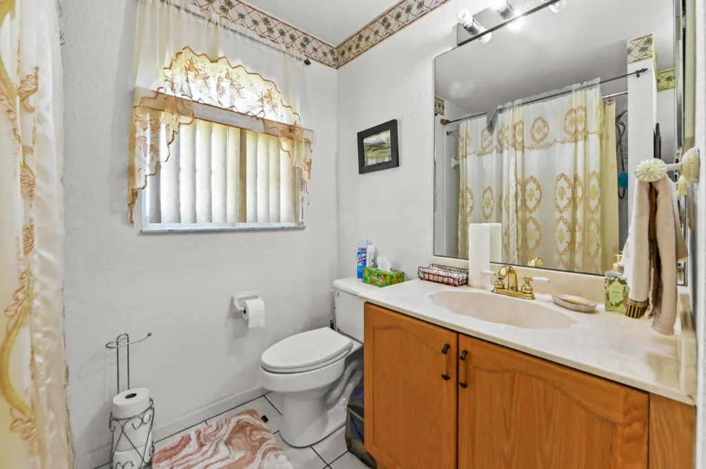 20261006193646505194000000-o - Lake Worth Homes - Lake Worth Homes Bathroom with white walls, gold-pattern shower curtain, window with vertical blinds, toilet, and light wood vanity with marble countertop.