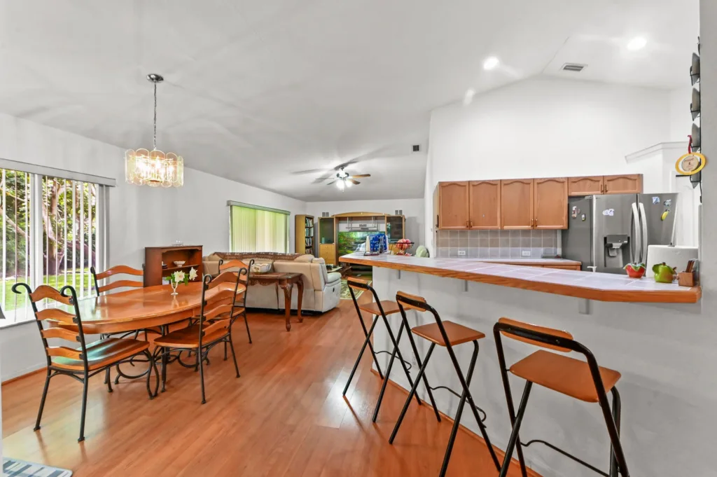 20261006193532303512000000-o - Lake Worth Homes - Lake Worth Homes Open-concept kitchen, dining, and living area with a wooden dining table, chairs, and a bar-height island with stools in the foreground.