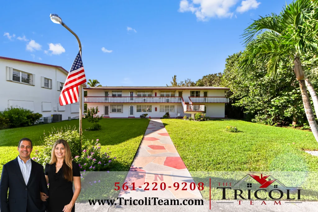 Two-story residential building with a flagpole and American flag in the front yard, palm trees, and two smiling real estate agents at the lower left.