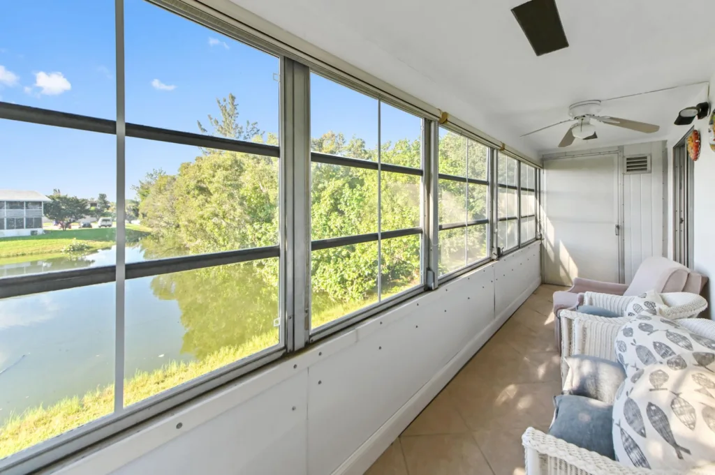Sunroom with large windows overlooking a pond and trees; white wicker chairs with cushions along the right side.