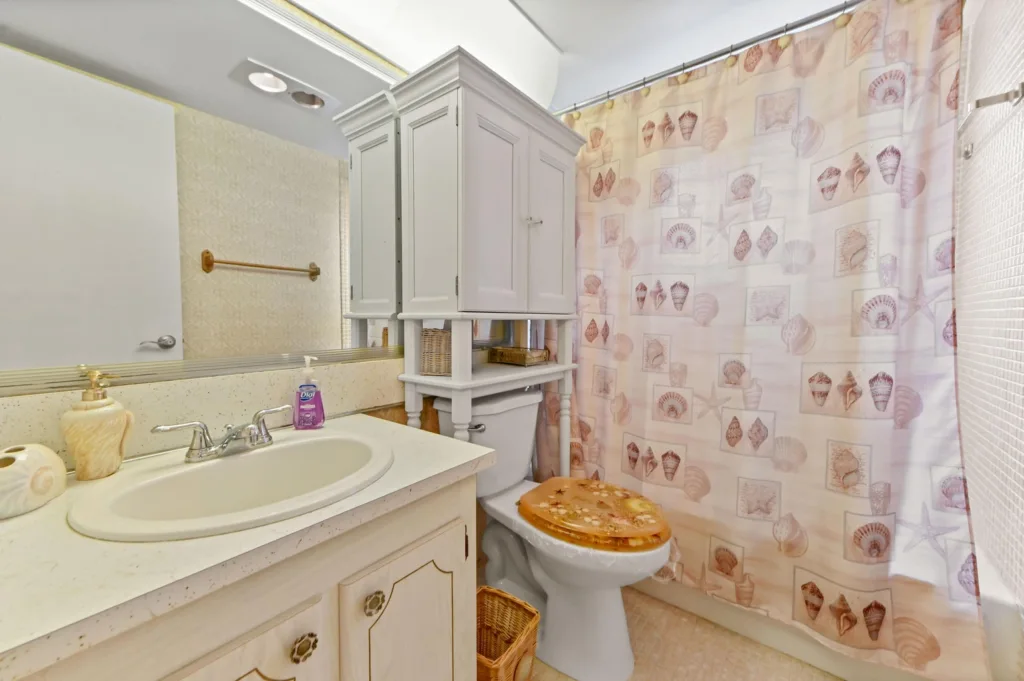 White bathroom with a vanity sink, countertop, brass faucet, and a large mirror overhead; a storage cabinet stands above the toilet.