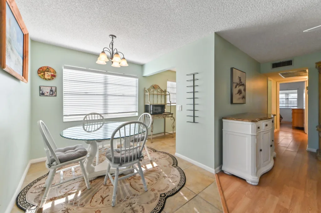 Dining area with a glass round table and white chairs on a floral rug; mint walls and blinds at the window.