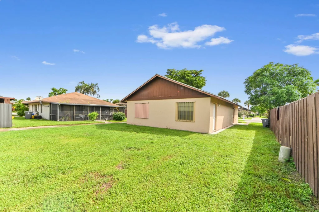 20261008162114551090000000-o - Lake Worth Homes - Lake Worth Homes Backyard scene with a beige, single-story house, brown gable roof, and a tall wooden fence on the right under a blue sky.