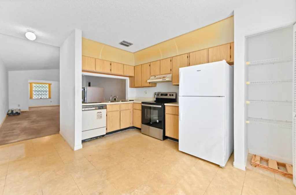 20261008161919803450000000-o - Lake Worth Homes - Lake Worth Homes Open-concept kitchen with wooden cabinets, beige tile floor, and white appliances (refrigerator, stove, dishwasher) arranged along the counters; leads to an adjacent living area.