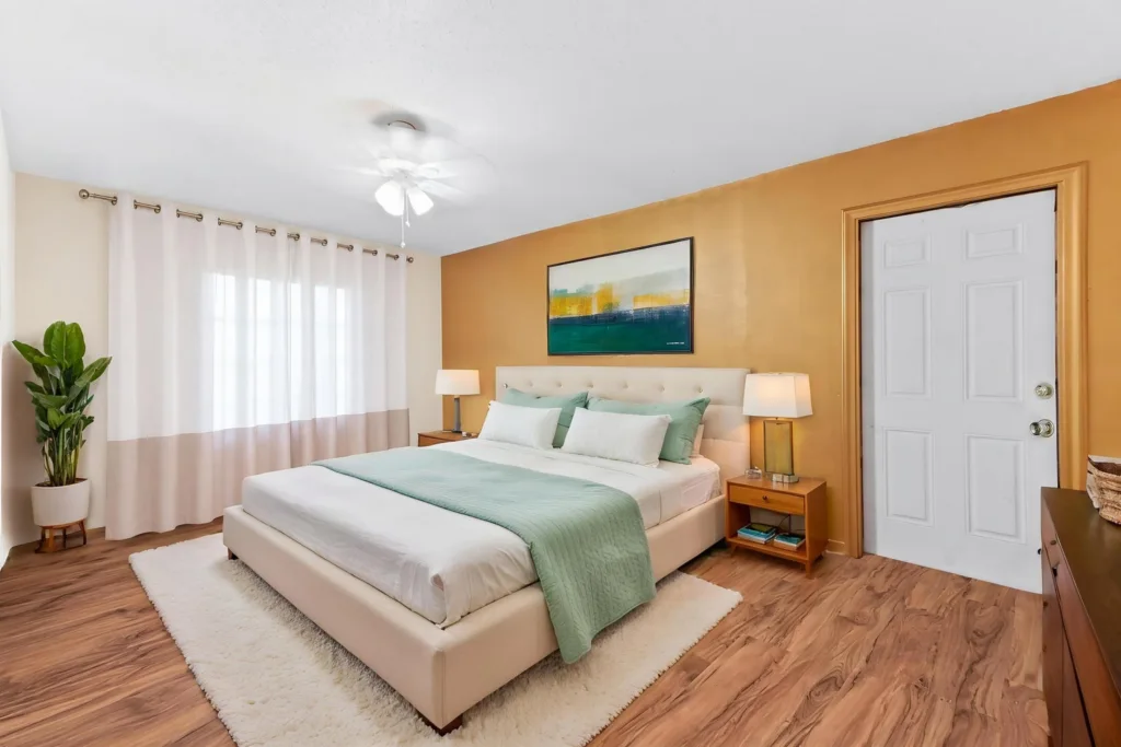 Bright modern bedroom with a beige upholstered king bed, teal bedding, and a white rug on wood floors under a warm orange accent wall.