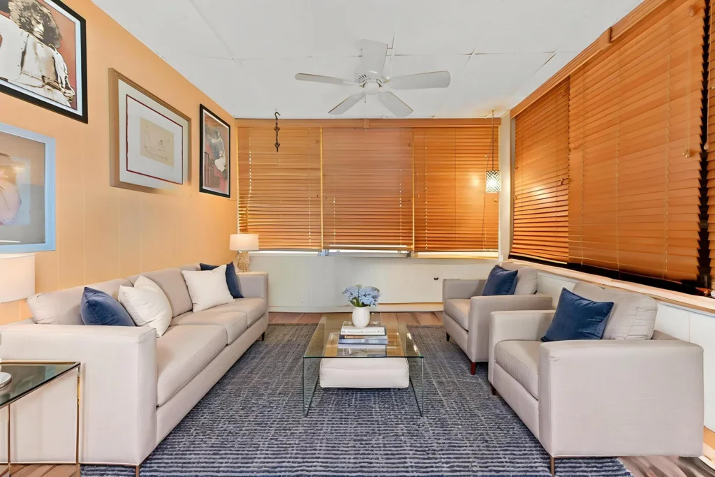 Modern living room with beige sofas, blue throw pillows, and a glass coffee table framed by orange blinds in the windows.