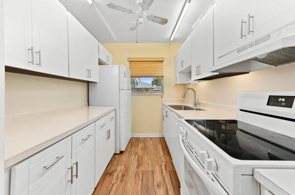Narrow white kitchen with upper and lower cabinets, white appliances, and a window at the far end. Hardwood floor runs along both sides.