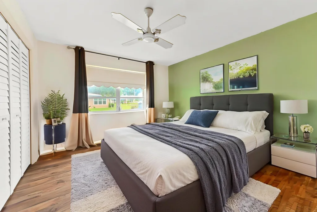 Bedroom with green accent wall and gray upholstered bed, white linens, navy throw, and two framed pictures above the headboard; matching nightstands and lamps on each side.